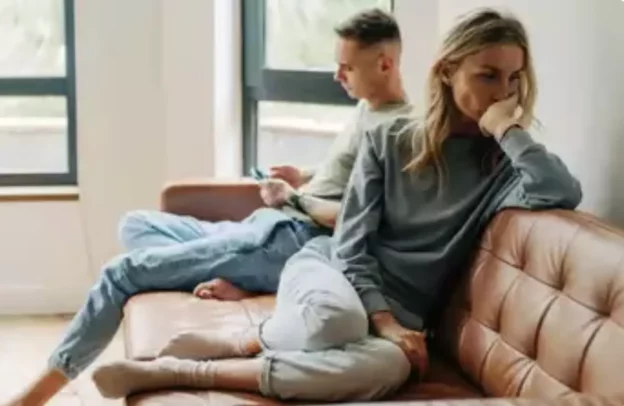 Woman on a beige leather sofa looks pensive, while a man in the background uses a device by the window.